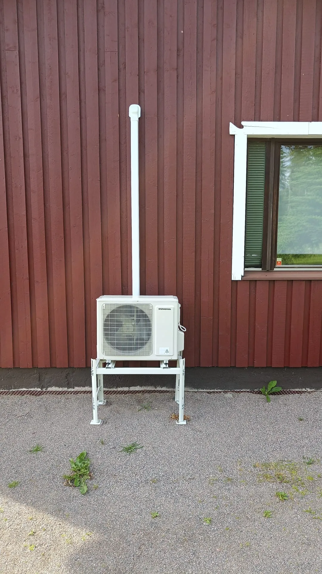 Punaruskeaa pystylautaseinää vasten on valkoinen Innova-merkkinen ilmalämpöpumpun ulkoyksikkö, joka seisoo valkoisella metallitelineellä. Yksiköstä nousee valkoinen putki ylös seinää pitkin. Oikealla on valkokehyksinen ikkuna, jossa näkyy tummanvihreä kaihdin ja puiden heijastuksia. Maassa on harmaata soraa ja muutamia vihreitä kasveja. Kuva on otettu kirkkaassa päivänvalossa.