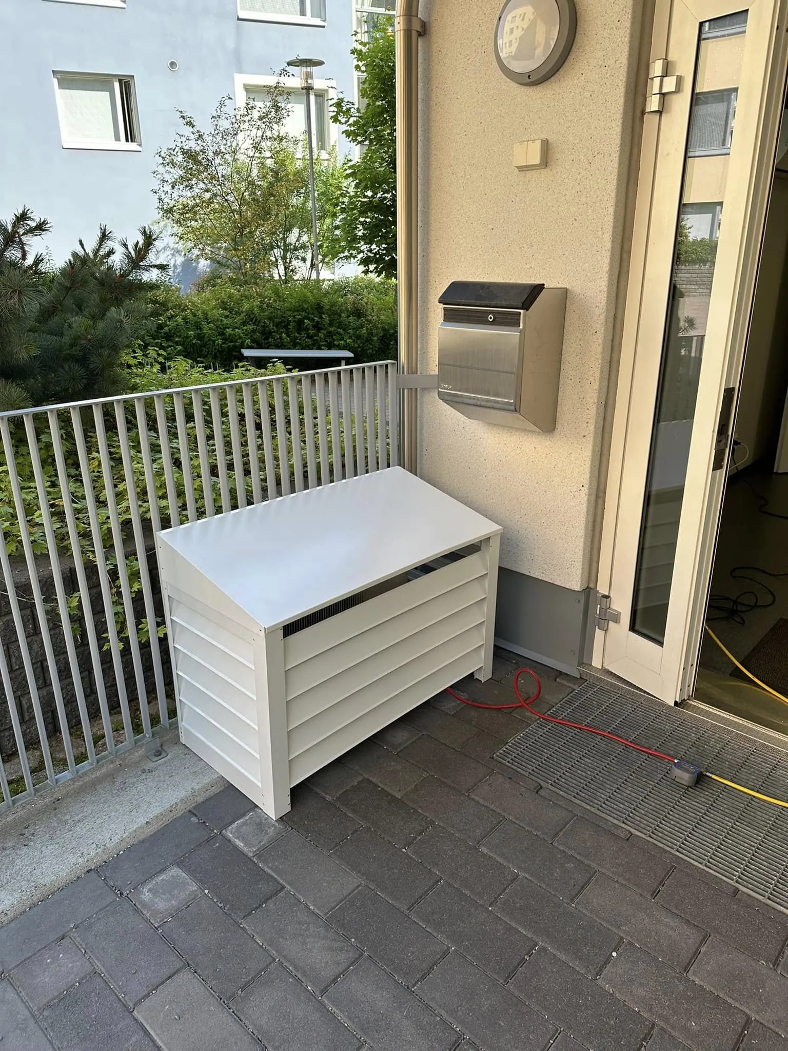 Päivänvalossa kuvattu ulkoterassi tai parveke. Vasemmalla on valkoinen, säleikköinen ulkosäilytyslaatikko viistokatolla. Sen vieressä on vaalea metallikaide, jonka takana näkyy tiheää kasvillisuutta ja taustalla vaaleansininen rakennus. Oikealla on rakennuksen vaalea seinä, jossa on ruostumattomasta teräksestä valmistettu postilaatikko ja pyöreä ulkovalo. Valokatkaisin on valon alapuolella. Seinän vieressä on hieman auki oleva valkoinen ovi. Maassa on tummanharmaita laattoja, metalliritilä sekä punainen ja keltainen johto.