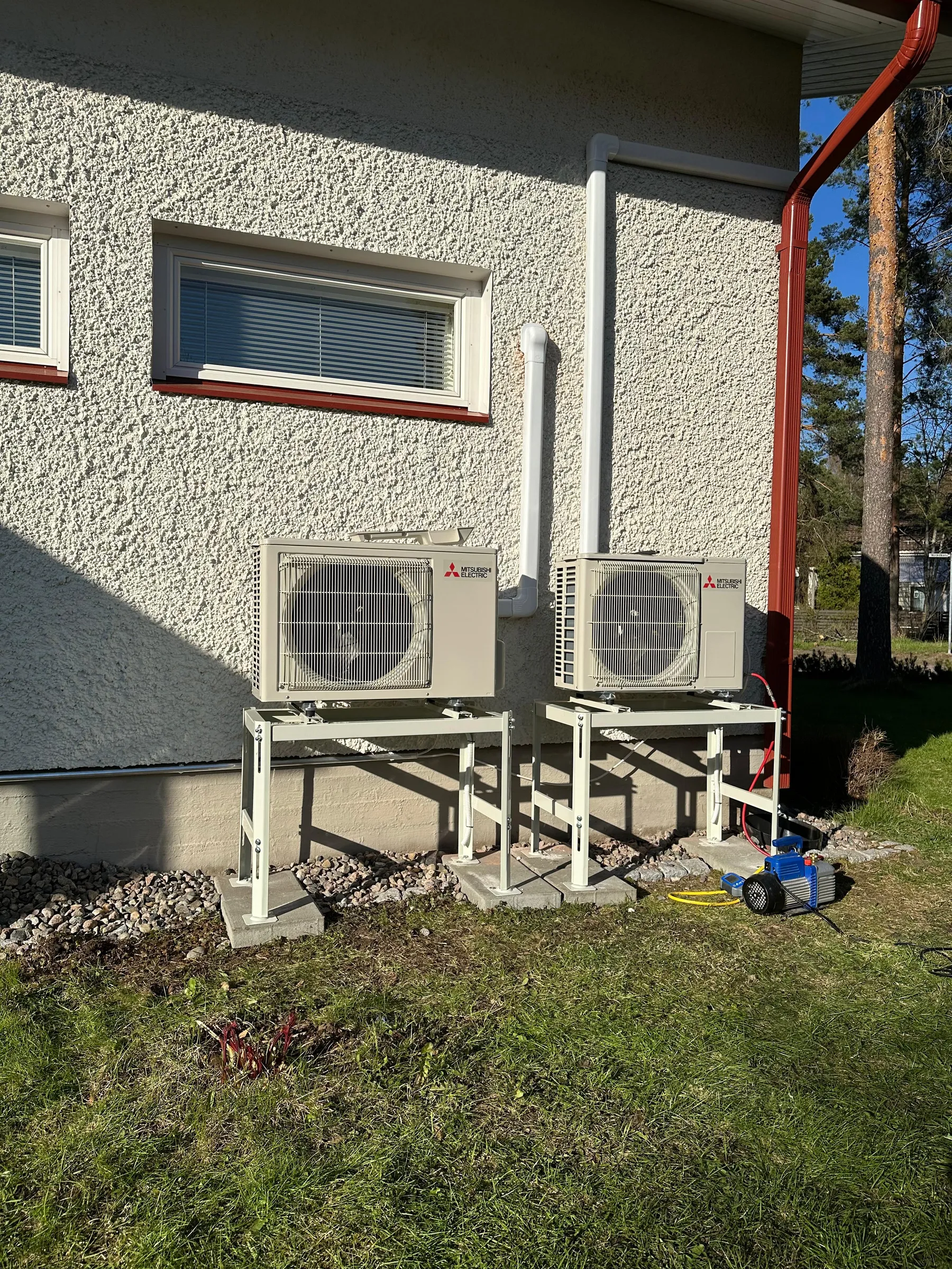 Kaksi Mitsubishi Electric -ulkoyksikköä seisoo valkoisilla telineillä talon rosoisen, vaalean seinän edessä. Seinällä on kaksi ikkunaa ja punaruskea syöksytorvi. Yksiköiden alla on nurmikkoa, soraa ja betonilaattoja. Oikealla nurmikolla on sininen ja musta tyhjiöpumppu keltaisine letkuineen. Aurinkoinen päivä luo teräviä varjoja seinälle. Taustalla näkyy puita ja sininen taivas.