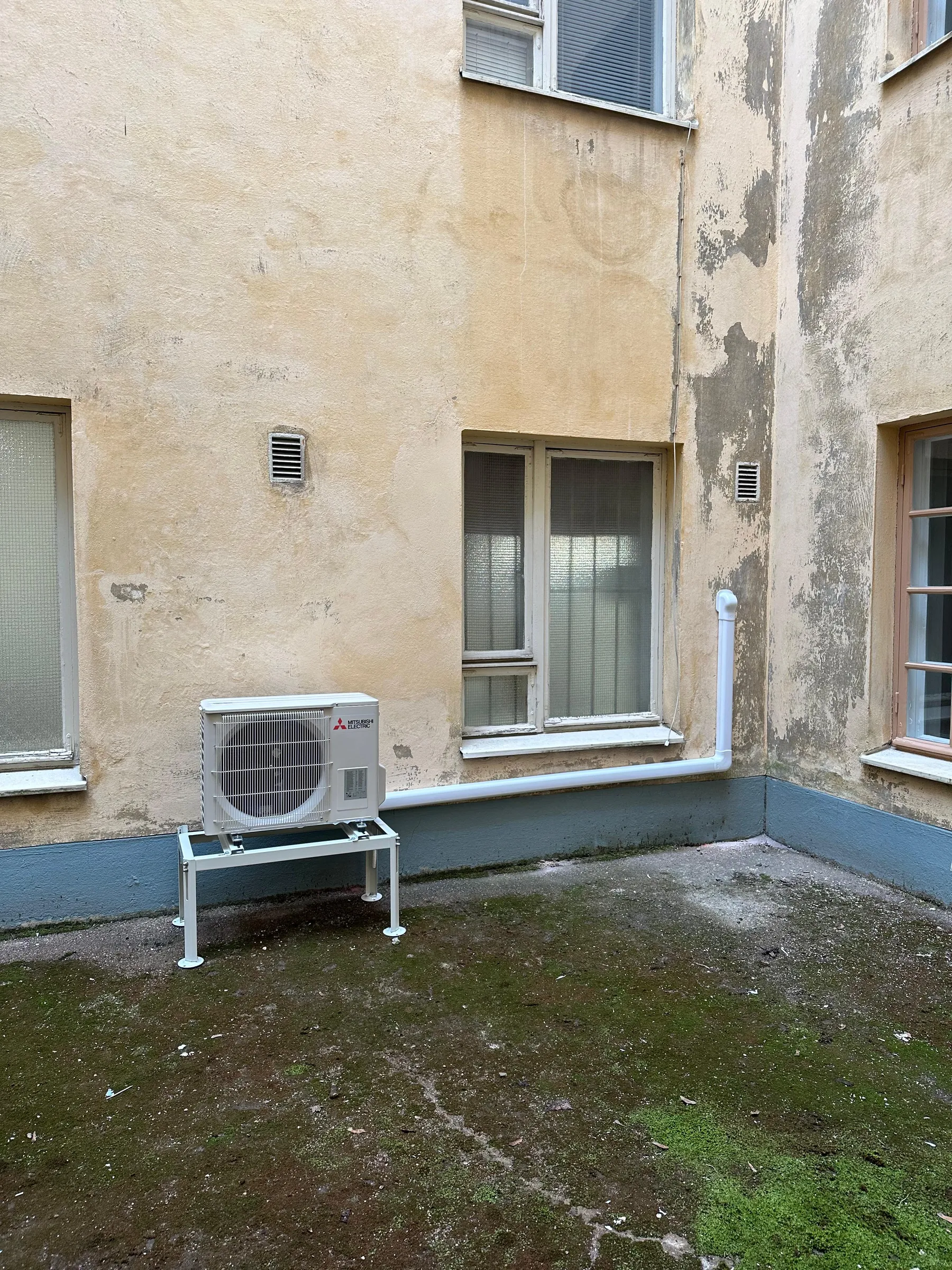 Ulkona oleva Mitsubishi Electric -ilmastointilaitteen ulkoyksikkö valkoisella telineellä. Se seisoo rapistuneen, vaaleankeltaisen rakennuksen seinää vasten, jonka alaosa on tummansininen. Seinässä on ikkunoita ja tuuletusaukkoja. Maanpinta on kostea ja sammalen peittämä.