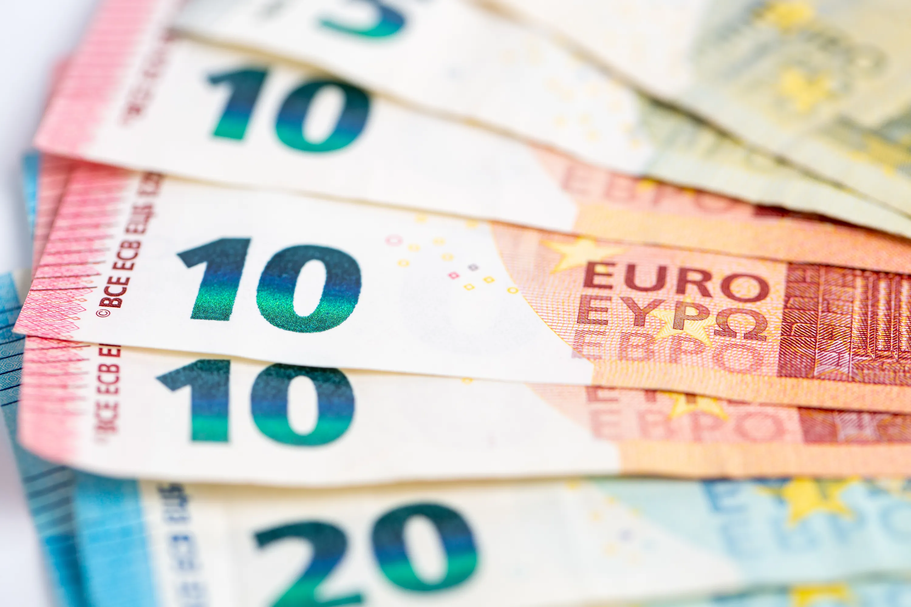 Lähikuva useista päällekkäisistä euroseteleistä valkoisella taustalla. Kaksi punaoranssia 10 euron seteliä on etualalla terävästi kuvattuna, niiden vihreänsiniset numerot '10' erottuvat selvästi. Alimpana näkyy osittain sininen 20 euron seteli. Ylimmät setelit ovat sumeita.