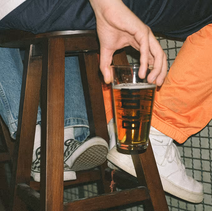 Hand holding a pint glass of beer near two people sitting on wooden stools with casual shoes. (Yasin Aribuga, unsplash)