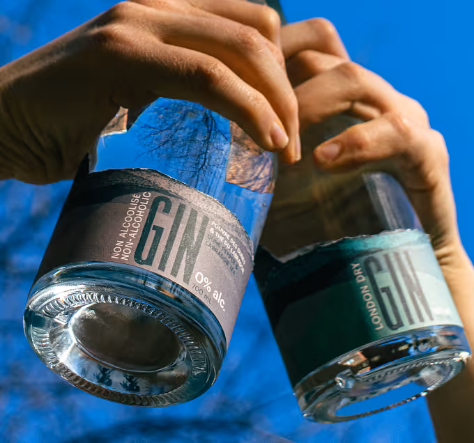Two hands holding bottles of non-alcoholic gin against a clear blue sky.