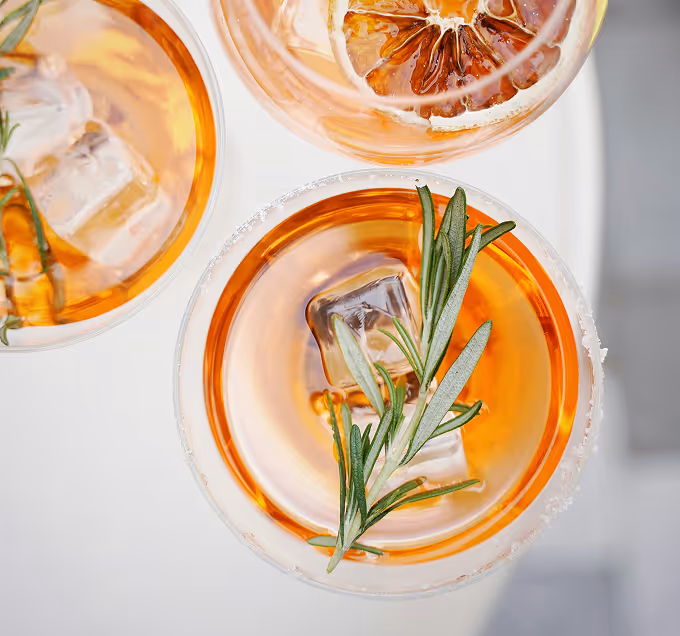 Overhead view of amber cocktails with ice, rosemary sprigs, and a dried citrus slice on a white table. (Olena Bohovyk, unsplash)
