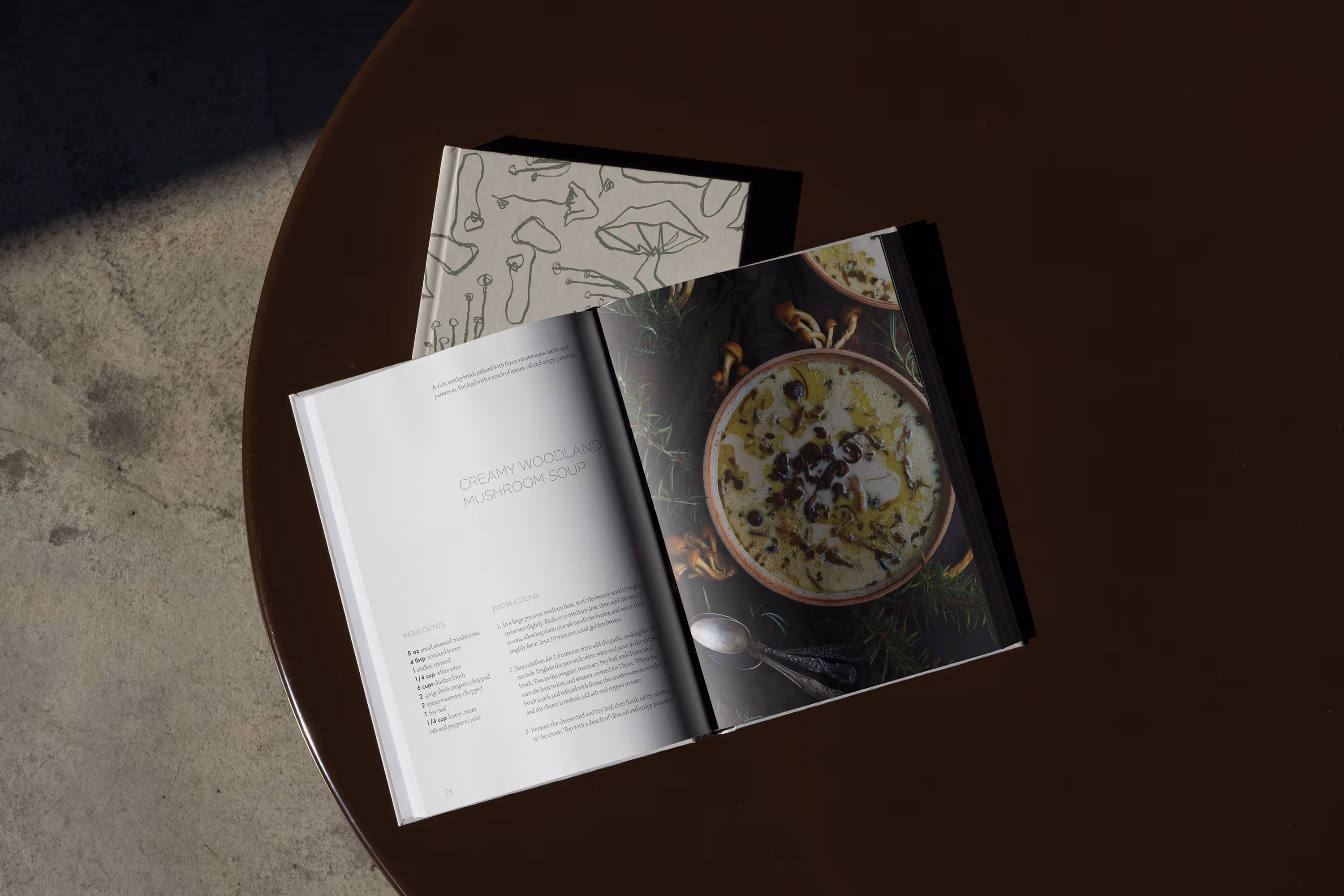 Open cookbook showing a recipe and image of creamy woodland mushroom soup on a dark table. mockup.maison)