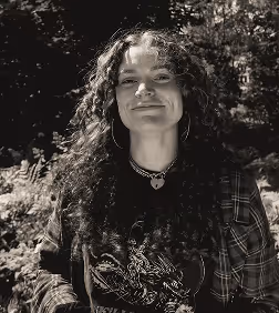 Smiling young woman with curly hair outdoors, wearing hoop earrings and layered necklaces including a heart pendant.