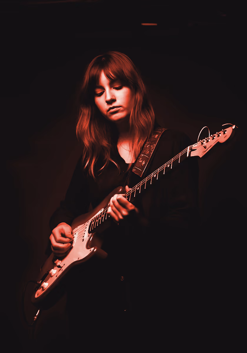 Young woman playing electric guitar, focused on the fretboard under red lighting
