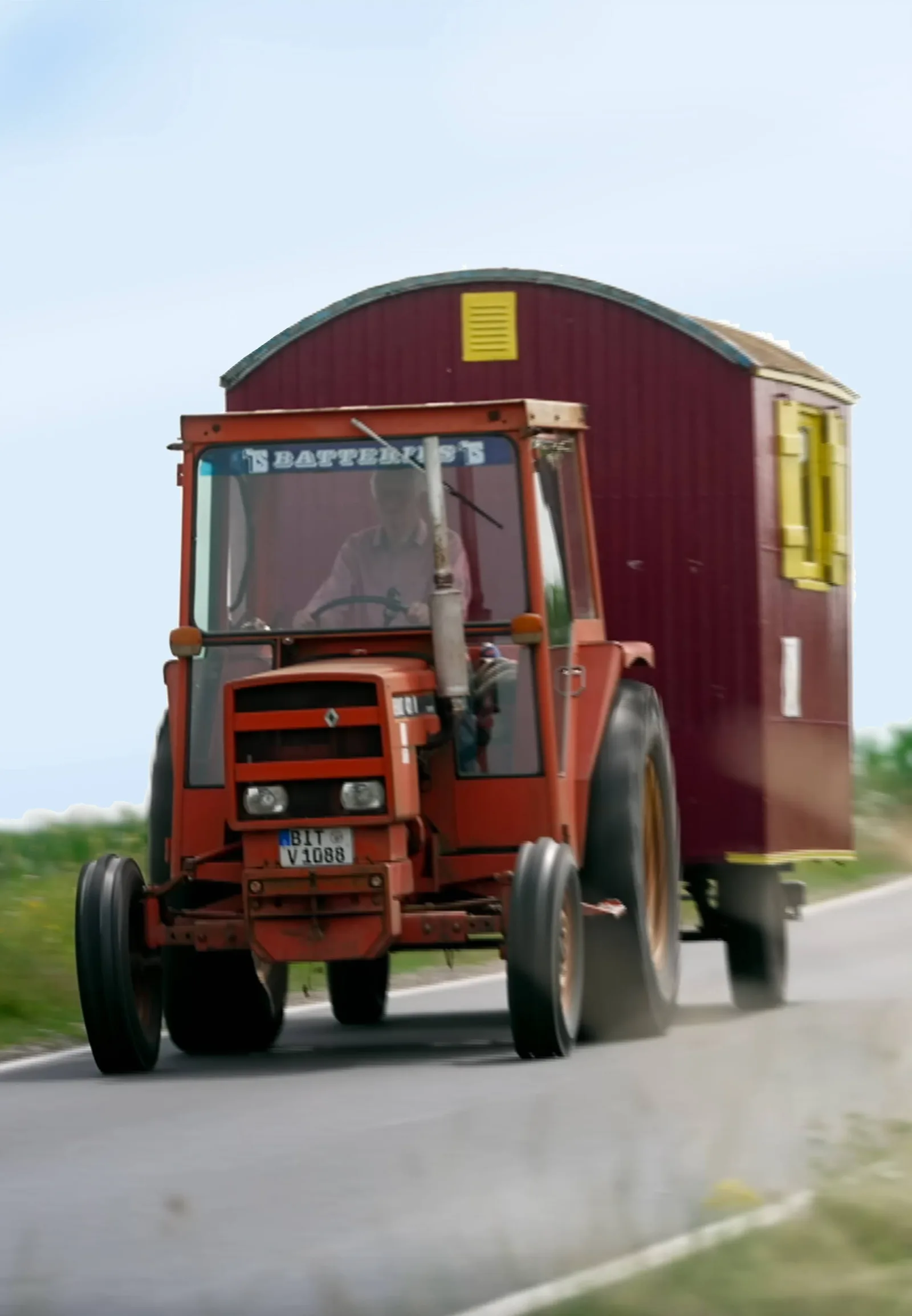 Roter Traktor mit Anhänger, der aussieht wie ein Bauwagen, fährt auf einer Landstraße.