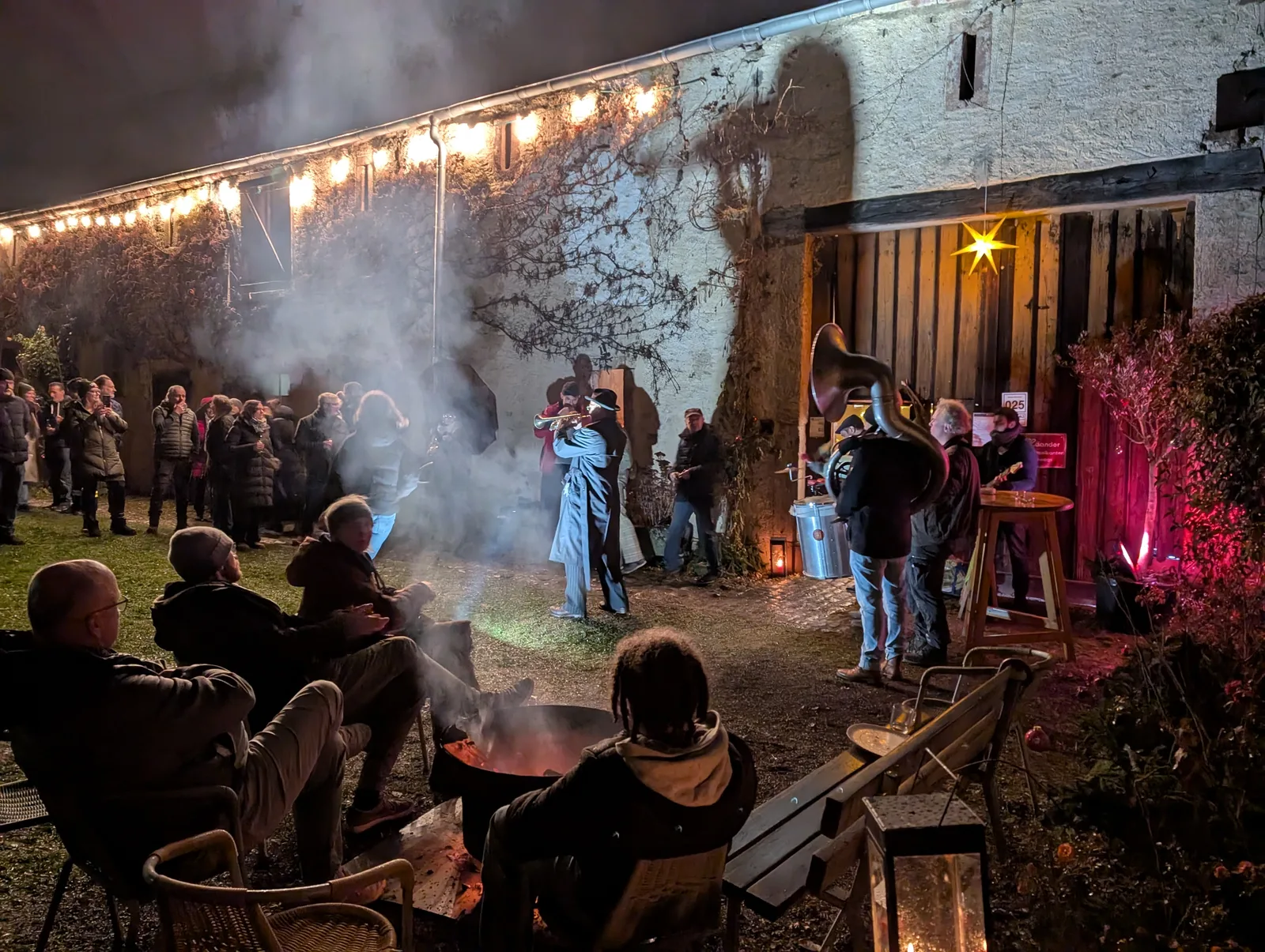 Menschen sitzen am Feuer, erleben Konzert mit Blasmusik vor beleuchtetem Haus bei Nacht.