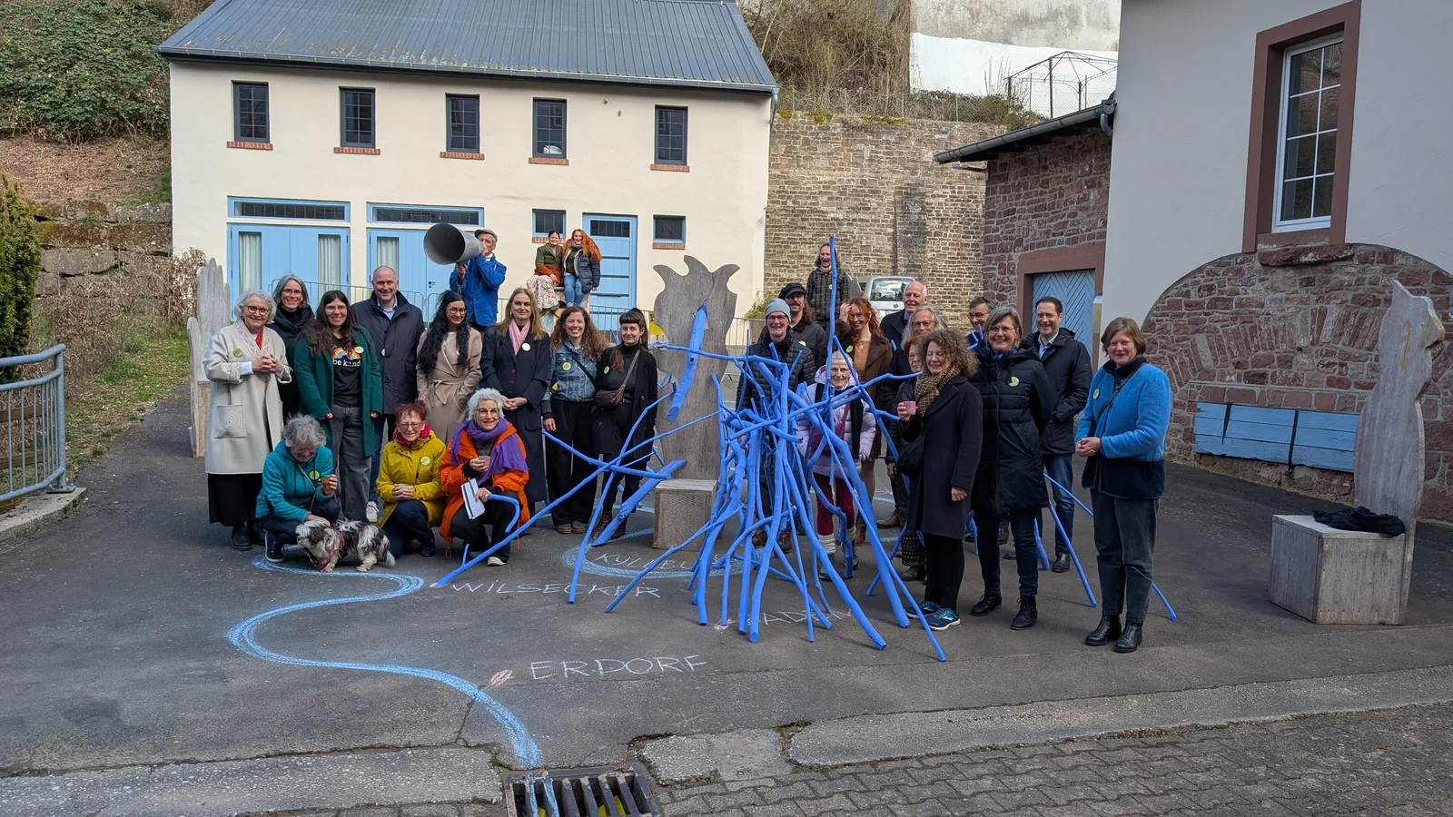 Gruppe von Menschen vor einem Gebäude mit blauen Türen und einer blauen Kunstinstallation aus Stöcken.