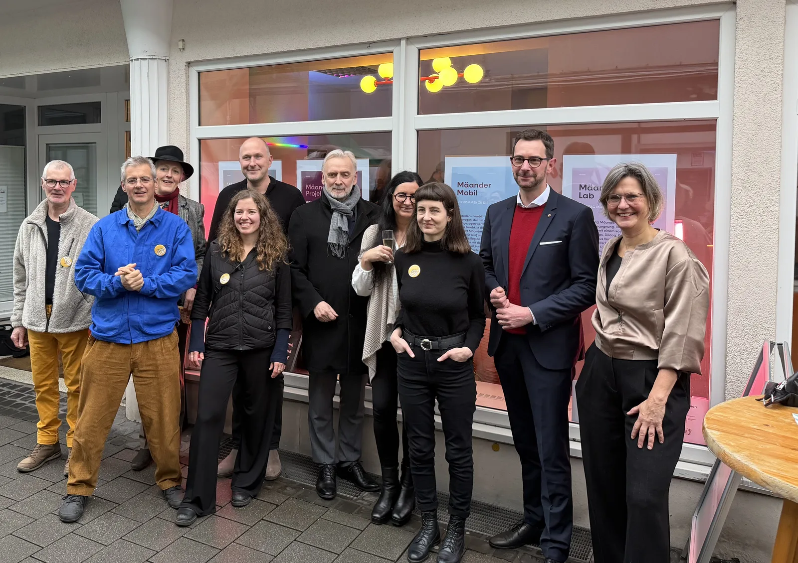 Gruppe von elf Personen steht vor einem Gebäude mit großen Fenstern und lächelt.