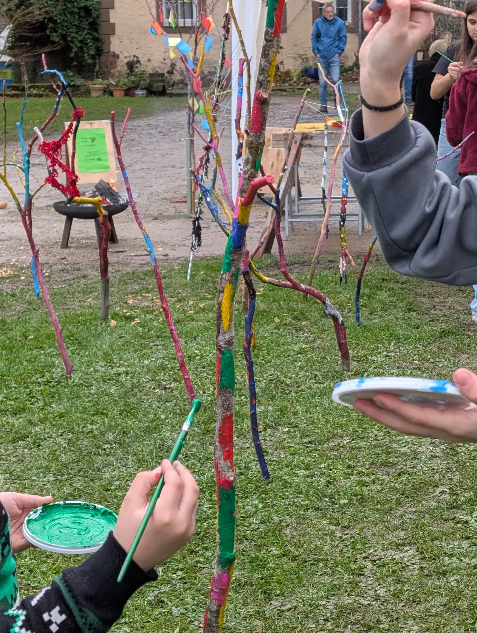 Menschen bemalen draußen bunte Äste, die in der Erde stecken, auf einer Wiese.