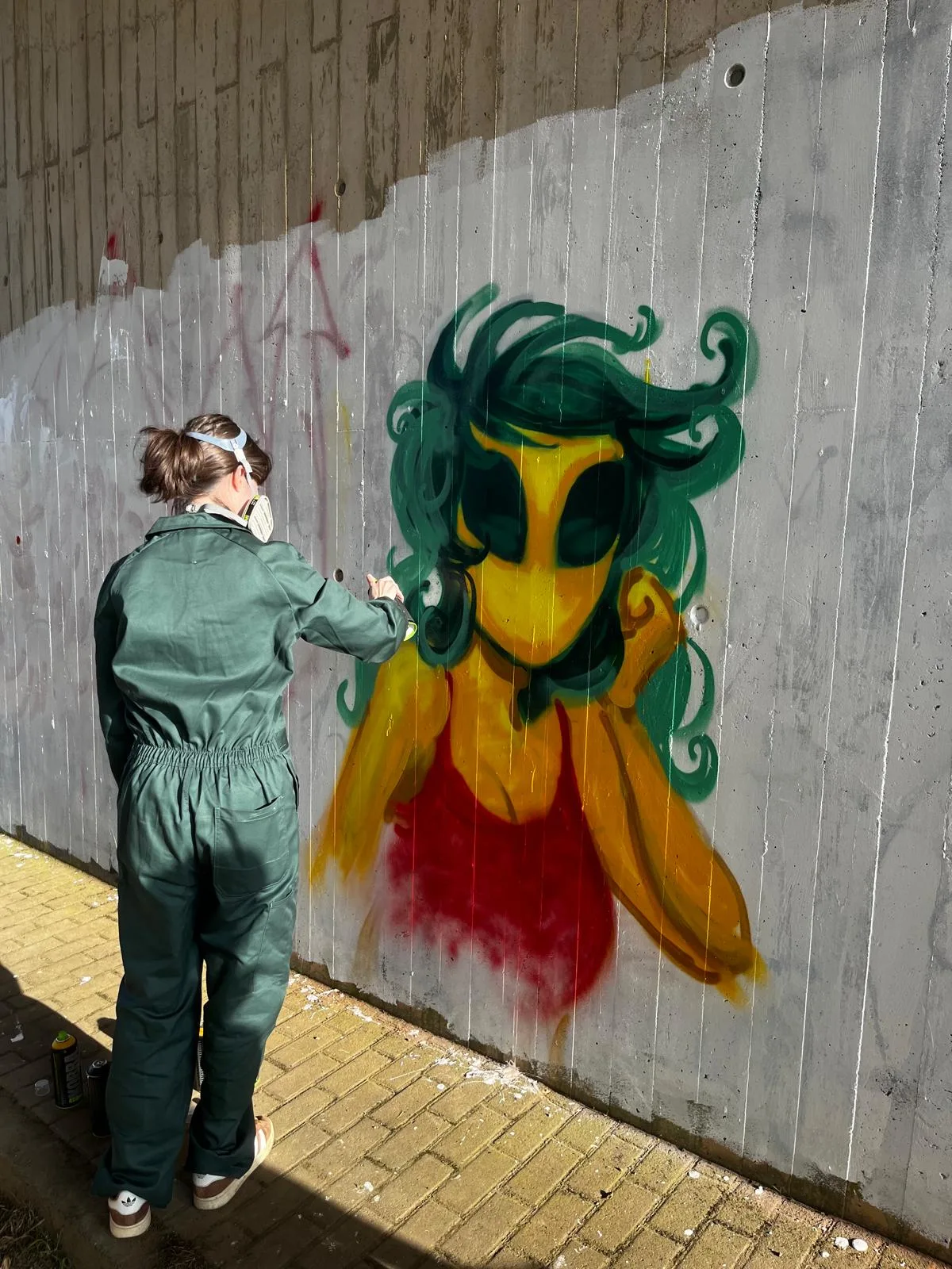Eine Person in grünem Overall sprüht Wandkunst mit einer gelben Figur mit grünen Haaren und rotem Oberteil.