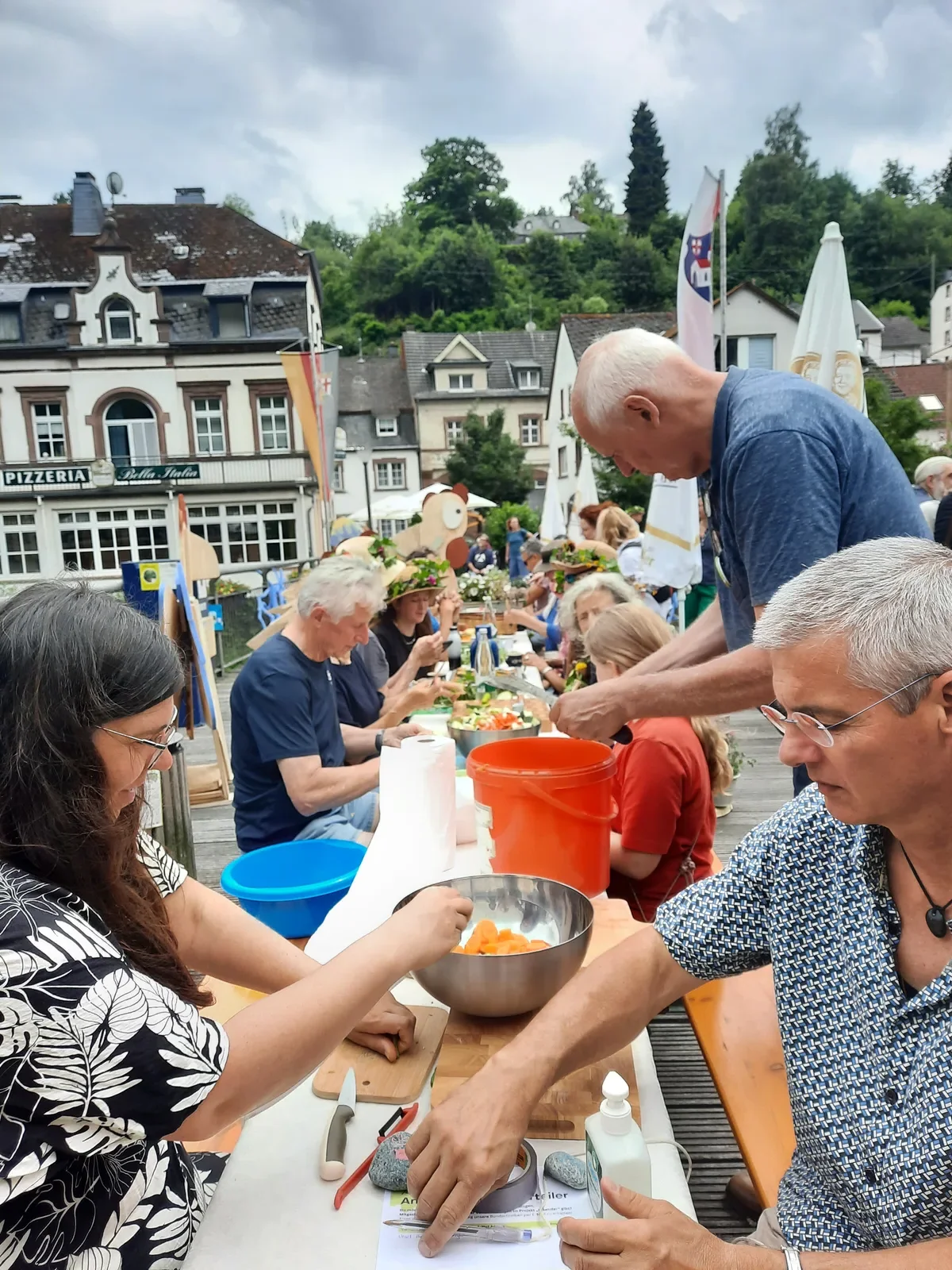 Menschen sitzen an einer langen Tafel im Freien und schneiden Gemüse bei bewölktem Himmel.