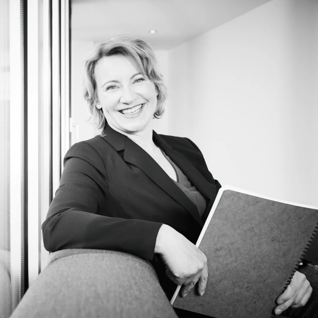 Frau sitzt lächelnd auf Sofa und hält einen Ordner im Arm in einem hellen Raum.