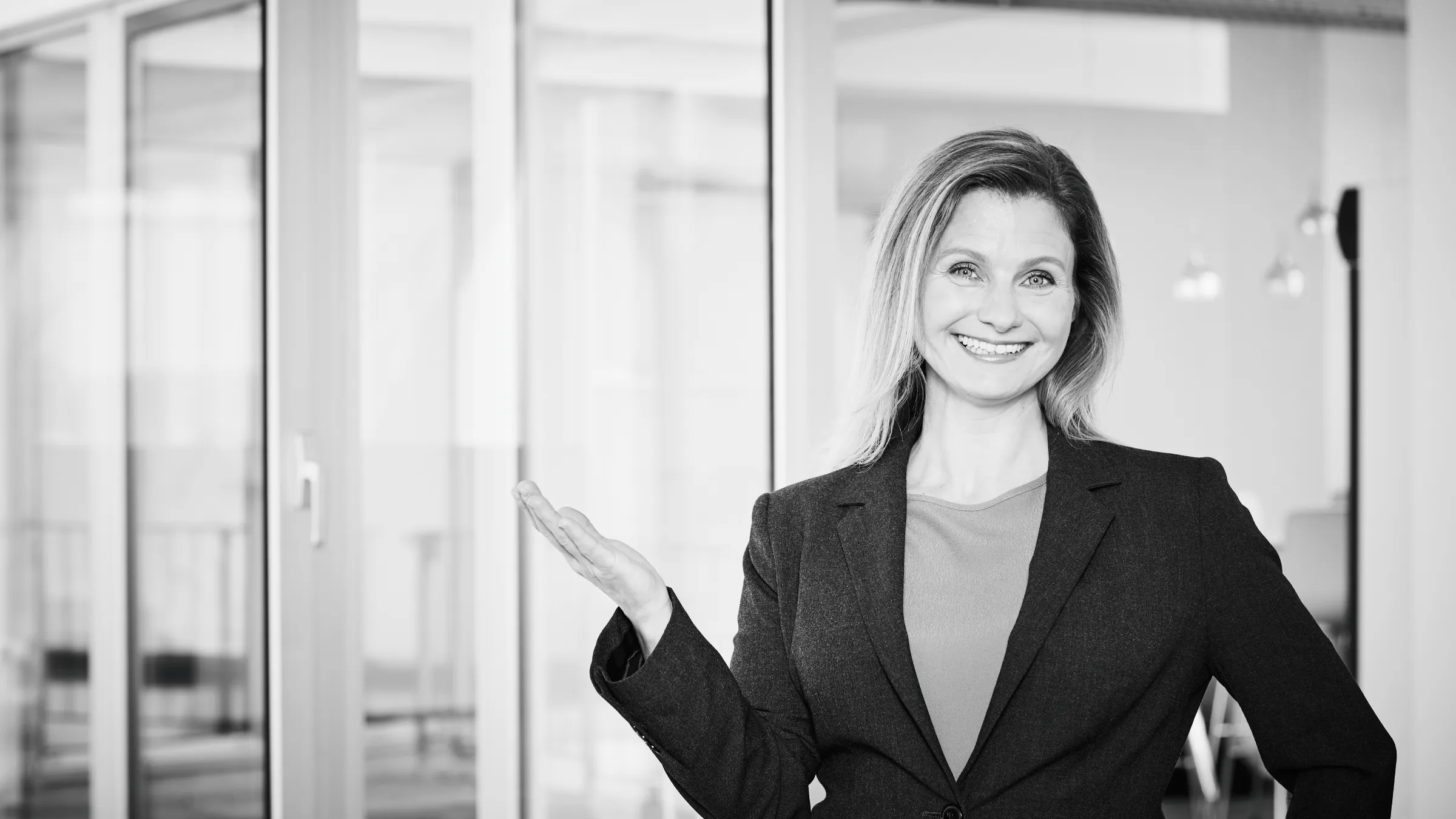 Frau im Businessanzug lächelt strahlend und hebt eine Hand in einem Büro mit Glaswänden.