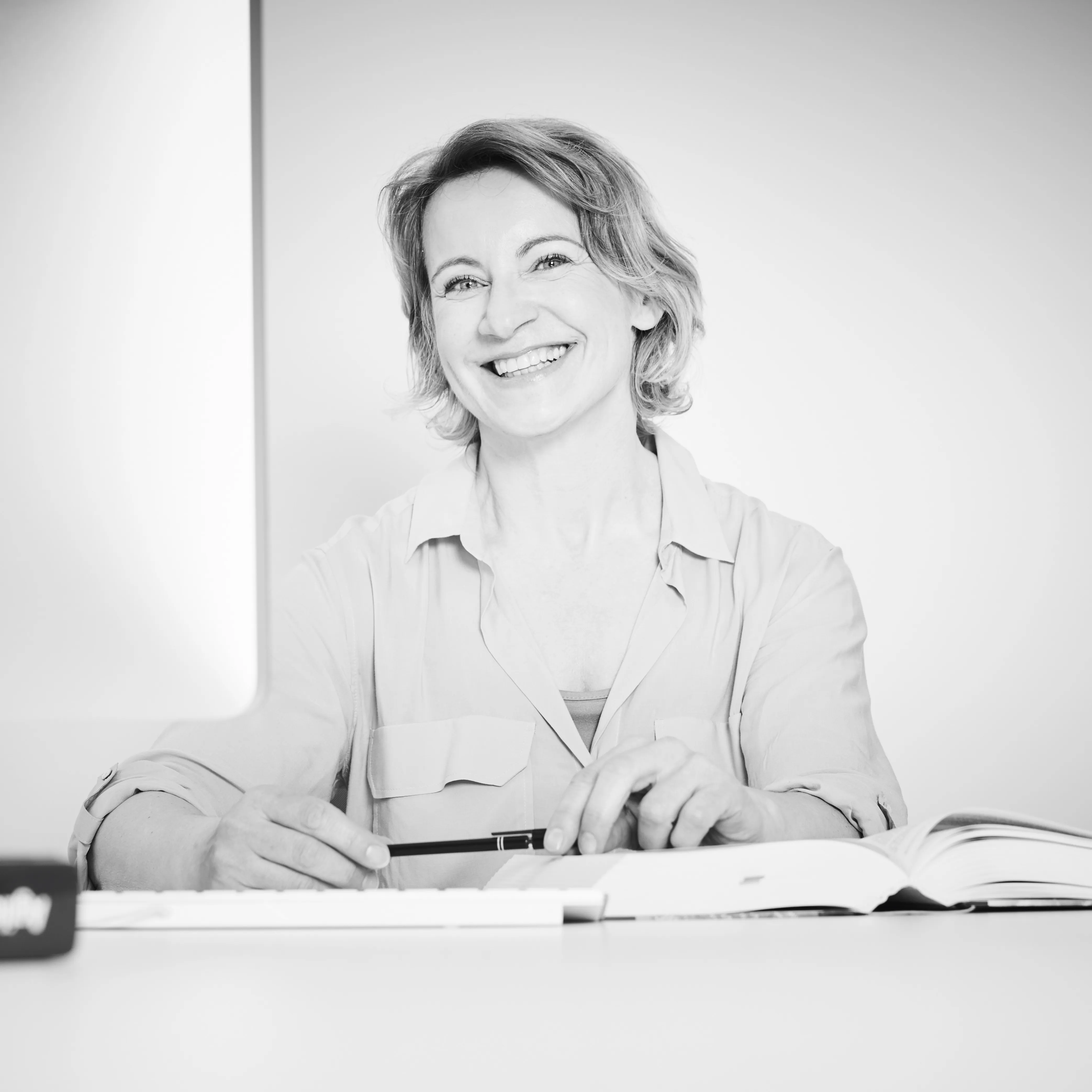 Lächelnde Frau sitzend an einem Schreibtisch mit aufgeschlagenem Buch und Stift in der Hand.