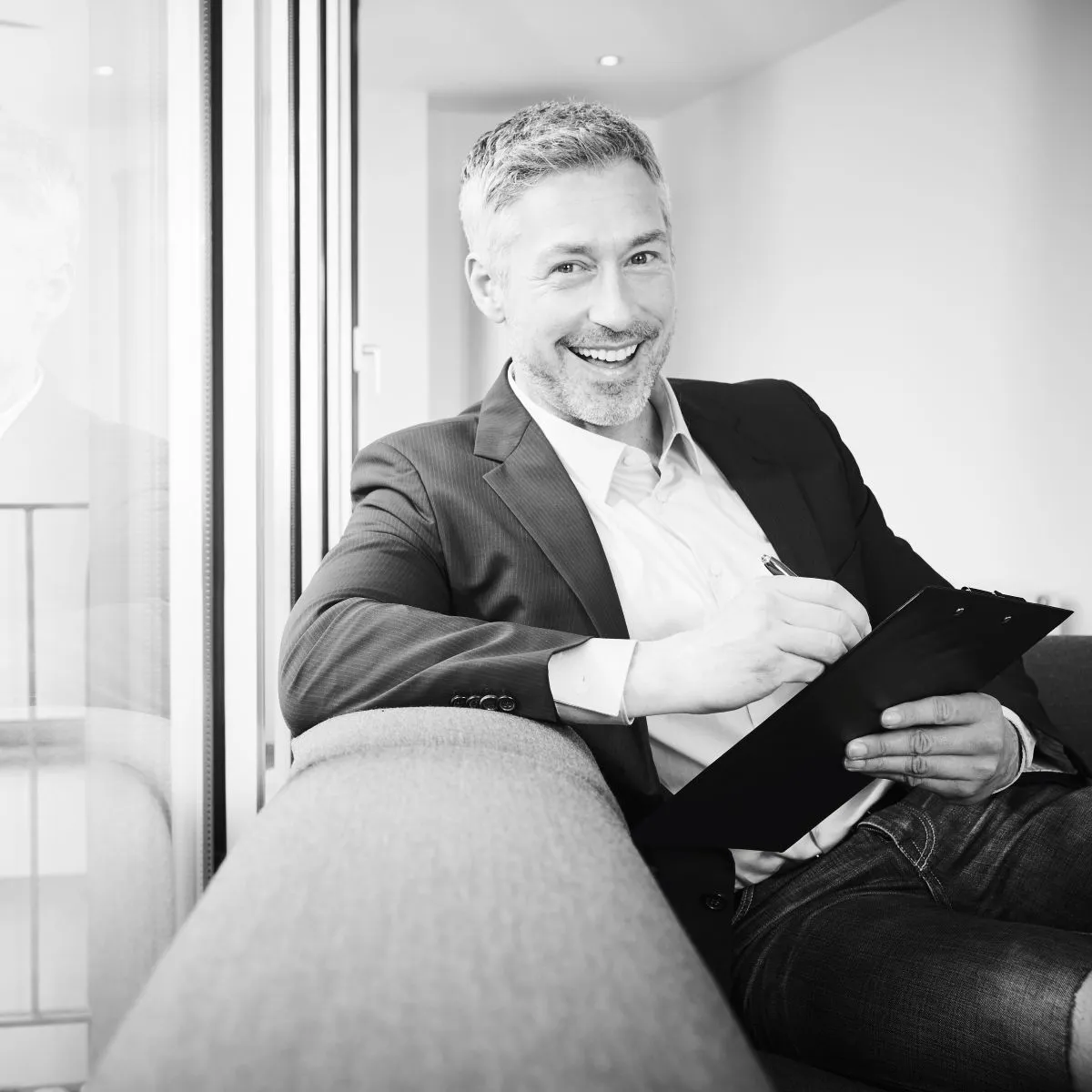 Lächelnder Mann in Anzug sitzt auf Sofa und hält Klemmbrett mit Stift.