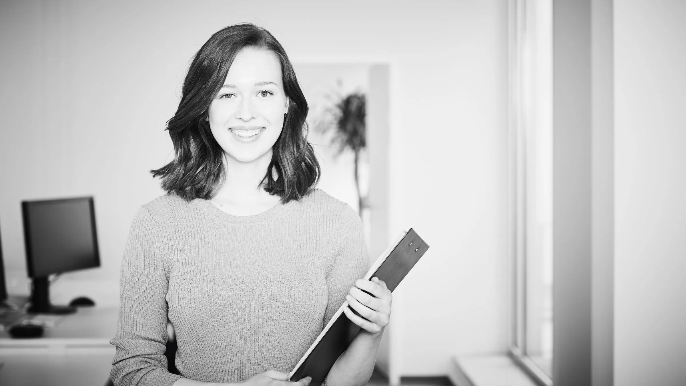 Lächelnde Frau im hellen Büro mit einem Klemmbrett in der Hand.