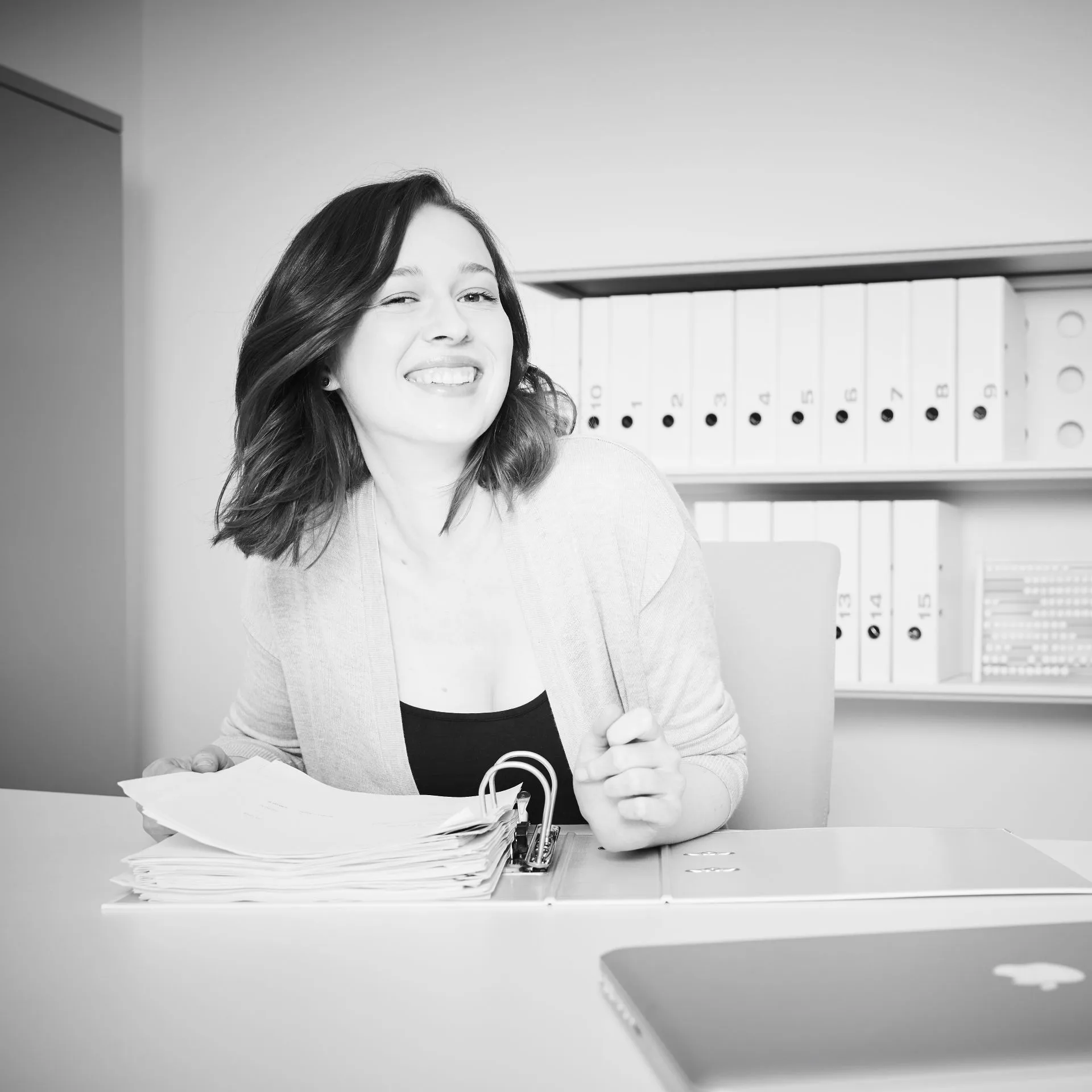 Lächelnde Frau sitzt am Schreibtisch mit offenem Ordner voller Papiere im Büro.