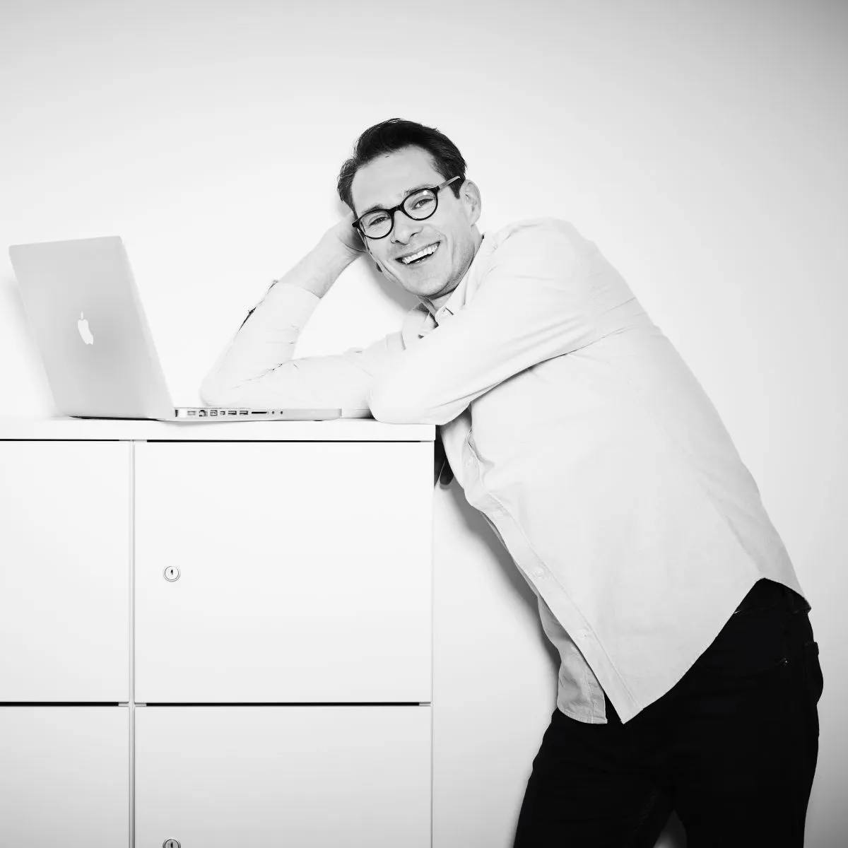 Lächelnder Mann mit Brille lehnt entspannt an Schrank neben offenem Laptop.
