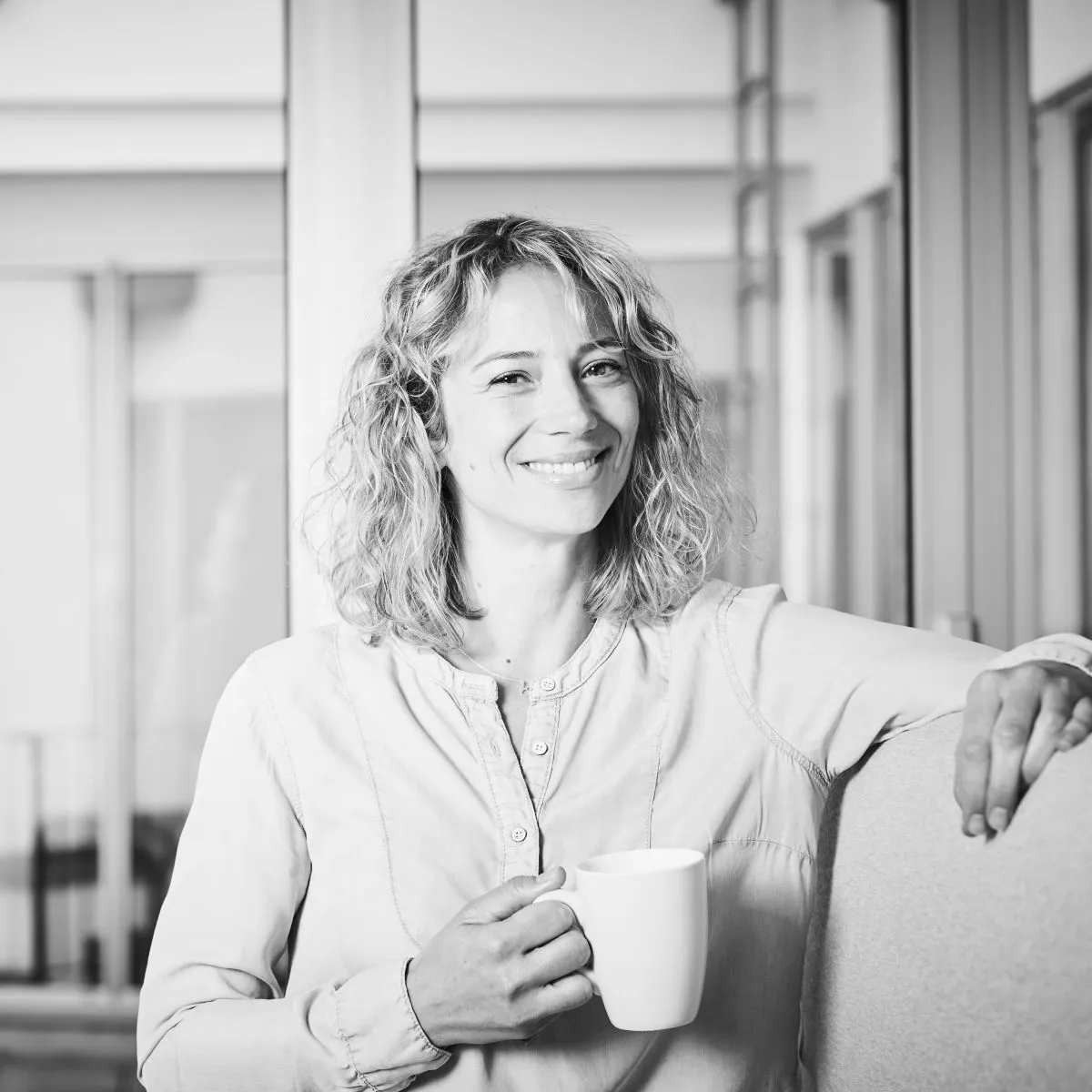 Frau sitzt lächelnd mit einer Tasse Kaffee auf Sofa in einem hellen Raum.