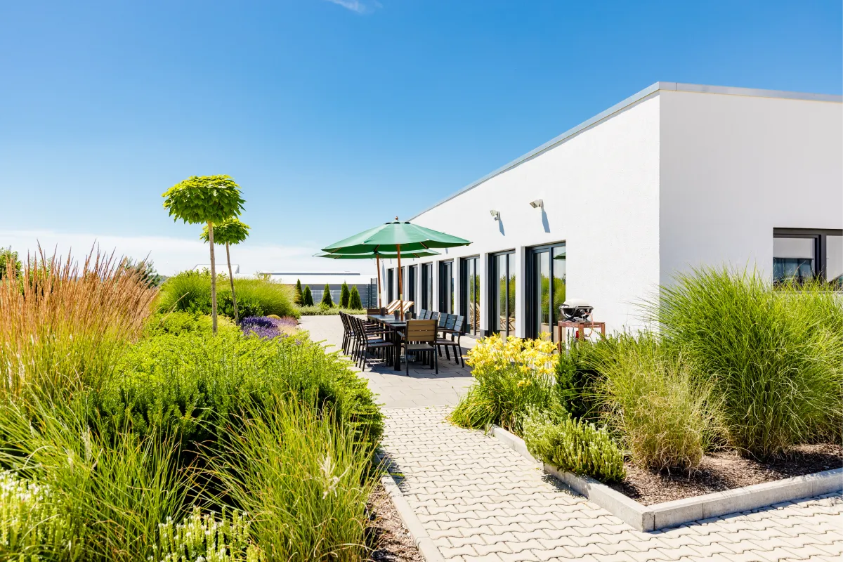 Terrasse mit Gartenmöbeln, grünem Sonnenschirm und gepflegten Pflanzen an modernem Gebäude.