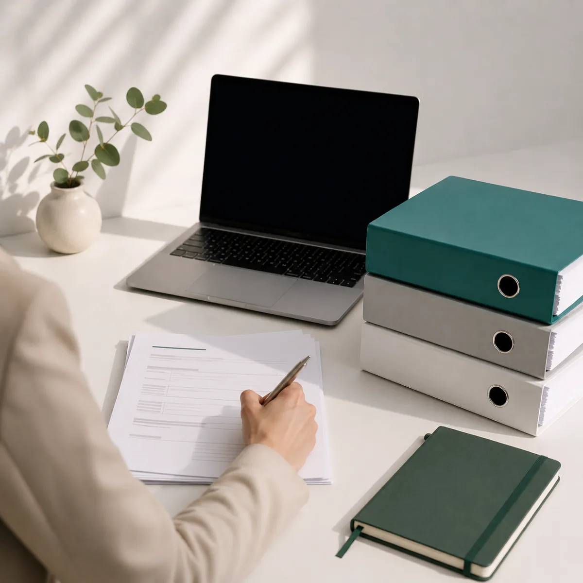 Person schreibt auf Papier am weißen Tisch mit Laptop, drei Ordnern, grünem Notizbuch und Pflanze.