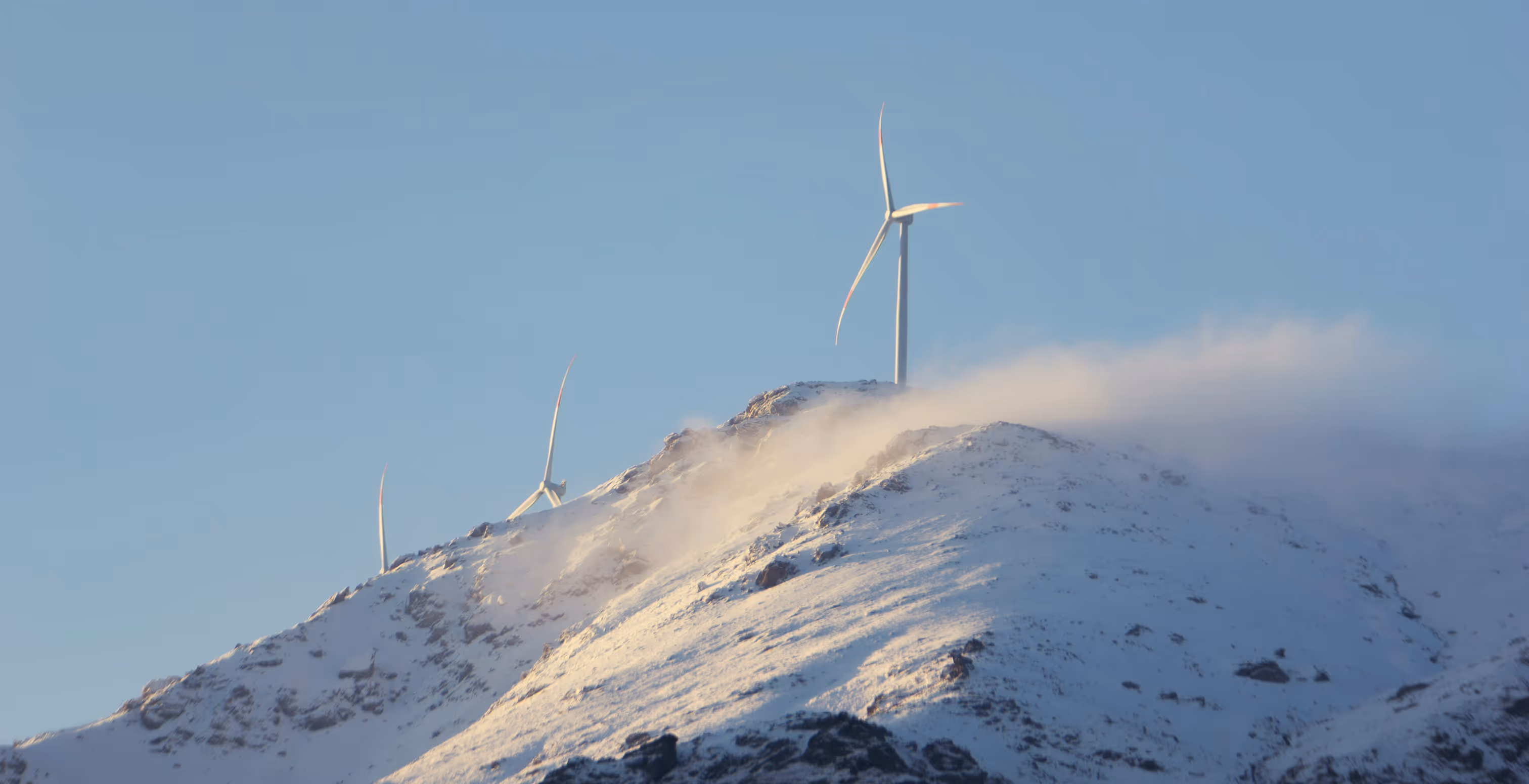 Wind turbines on a snow-covered mountain with mist and clear blue sky above.