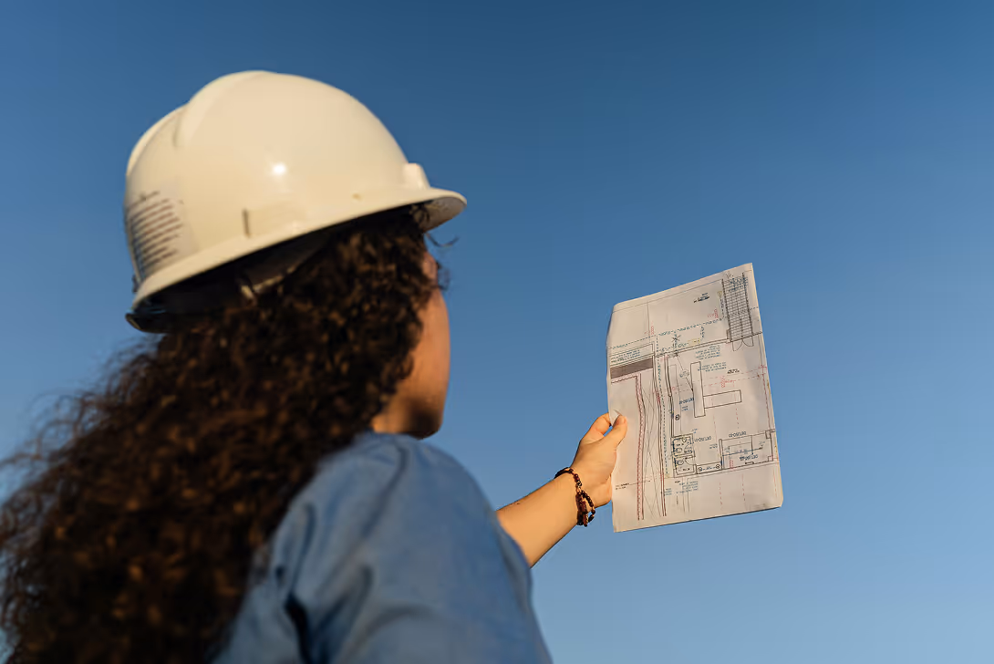 Person in white hard hat holding architectural blueprint against clear blue sky.