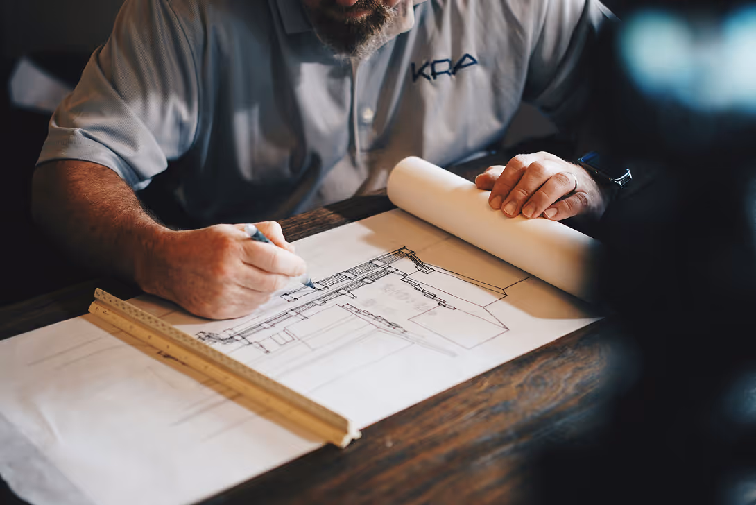 Man in gray shirt drawing architectural plans with ruler on wooden table.