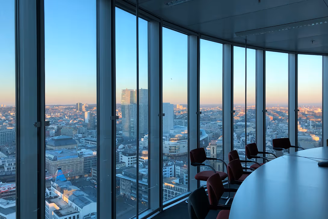 City skyline at sunset viewed through curved floor-to-ceiling windows of a conference room with chairs.