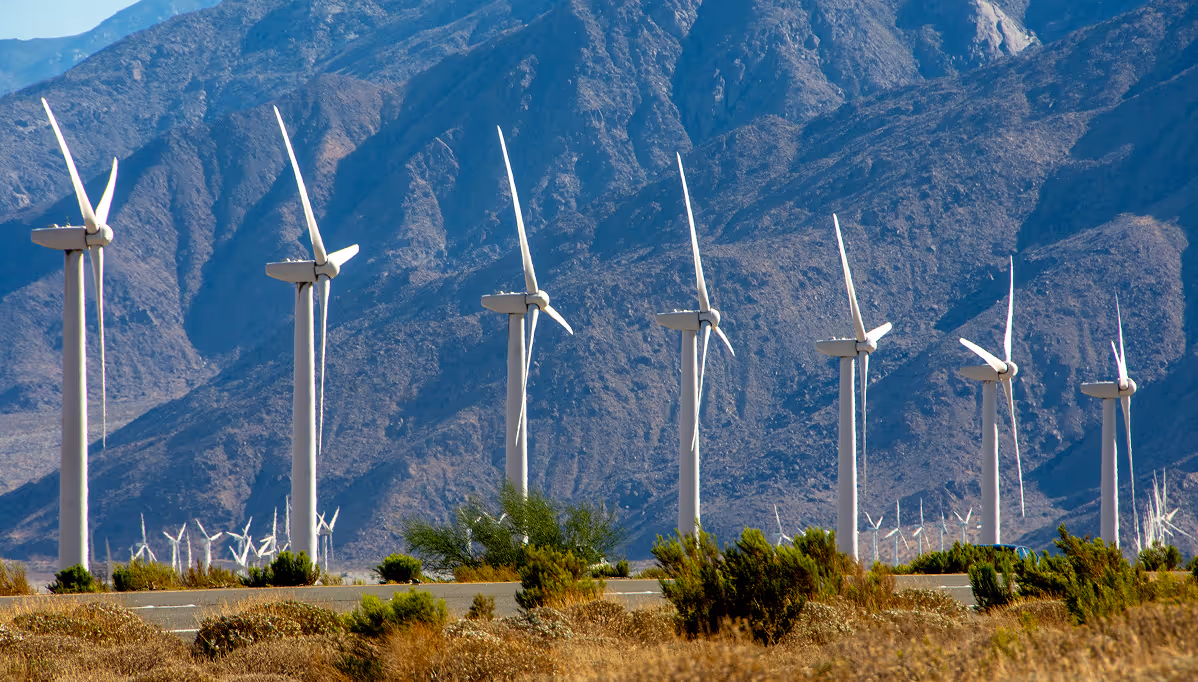 Row of wind turbines in a desert landscape with mountains in the background on a sunny day.