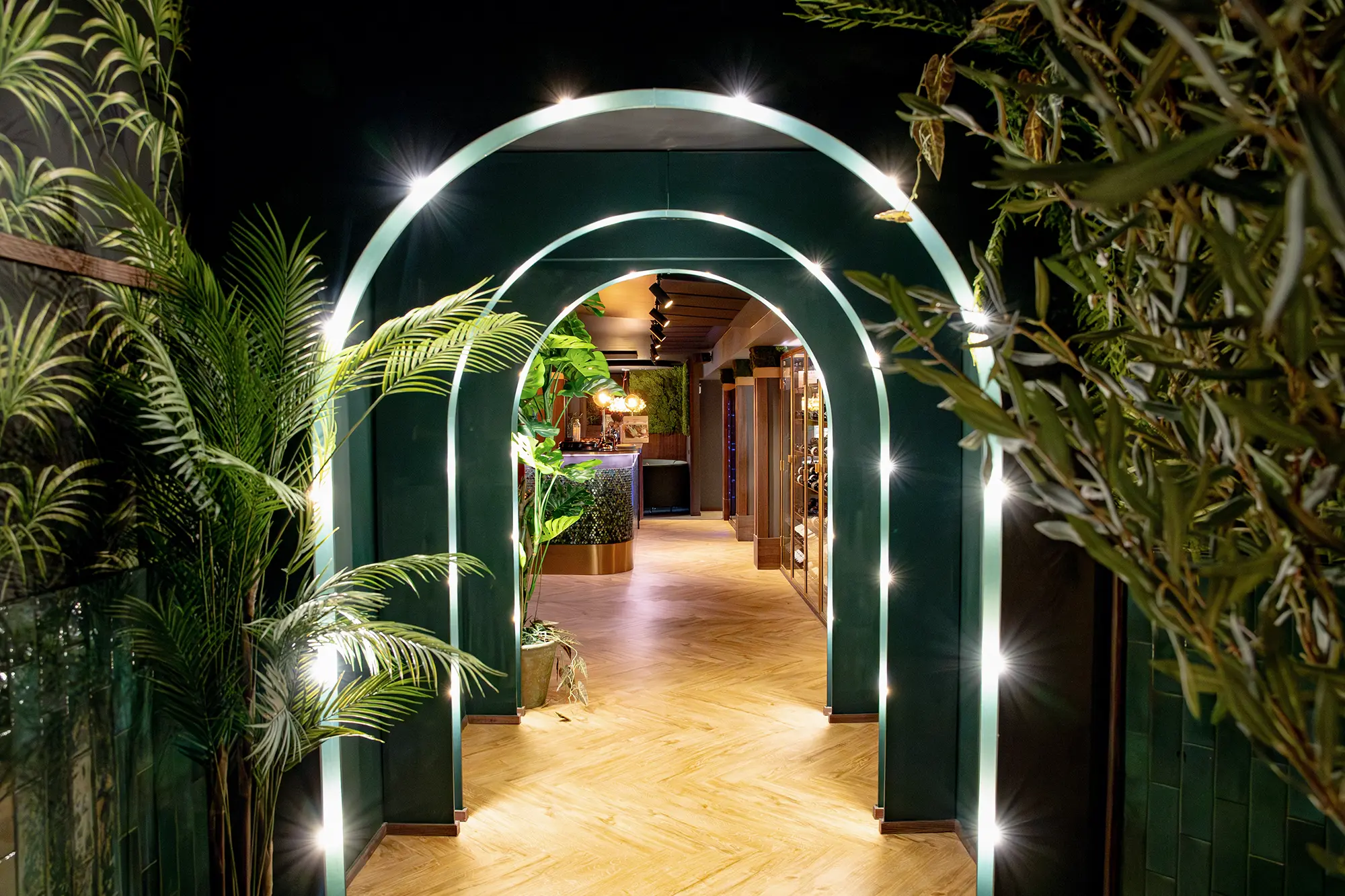 Indoor corridor with illuminated green arches, plants on sides, and wooden herringbone floor leading to a bar area.