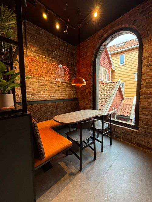 Cozy corner seating with orange bench, two chairs, wooden table, neon DIGG sign, and arched window view.
