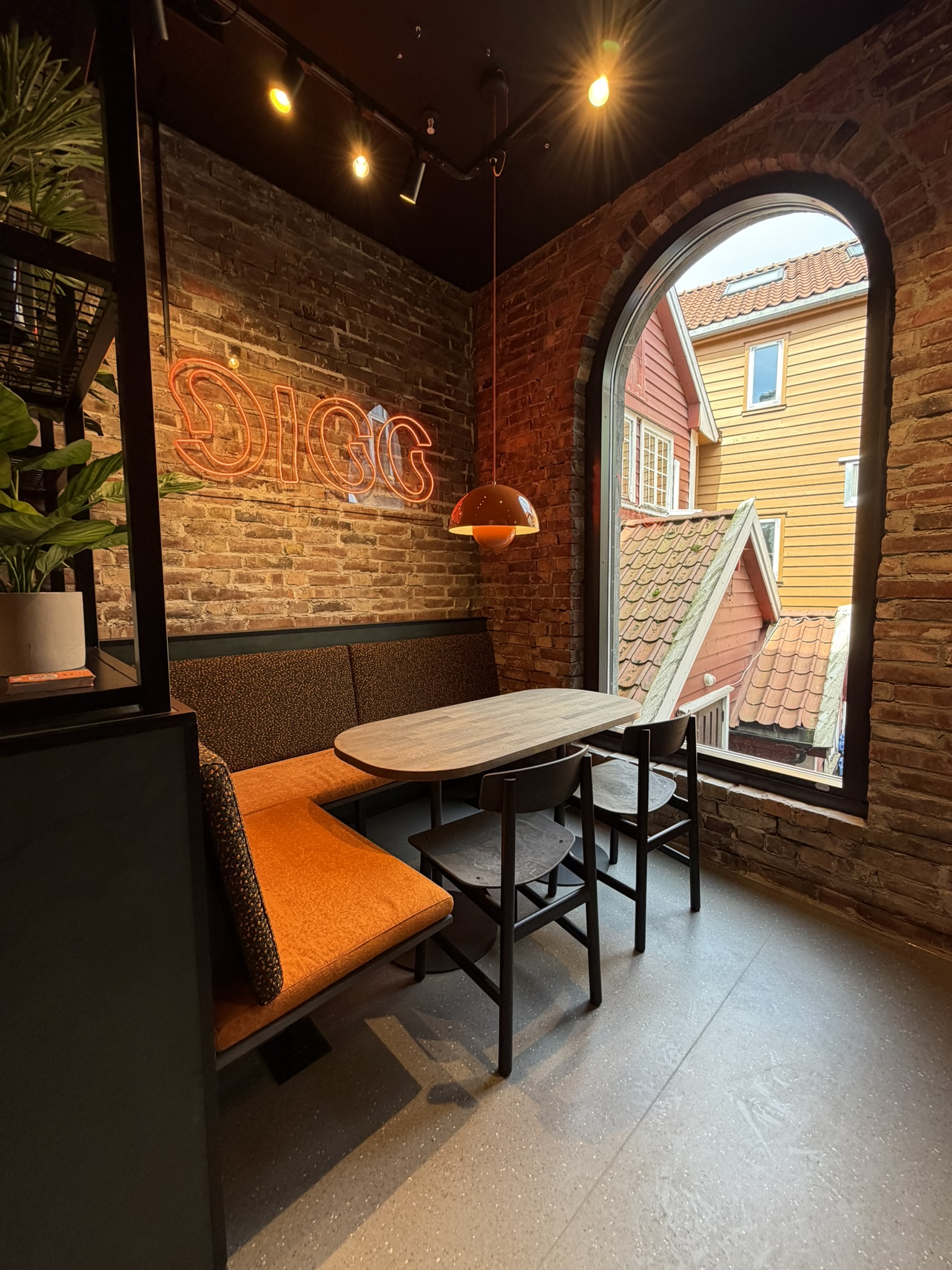 Cozy corner seating with orange bench, two chairs, wooden table, neon DIGG sign, and arched window view.