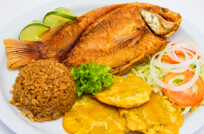 A plate of Dominican-style fried fish served with seasoned rice, crispy tostones, fresh salad, and a lime wedge.