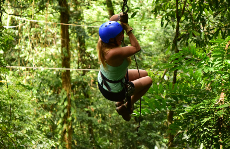 Zip-line rider flying above jungle canopy with sea view.