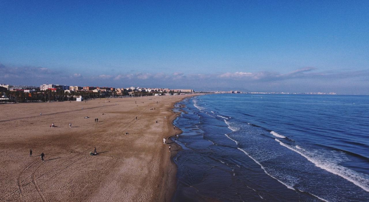 Wide sandy beach curving toward a coastal city skyline, with gentle waves and a few people walking along the shore under a clear blue sky.
