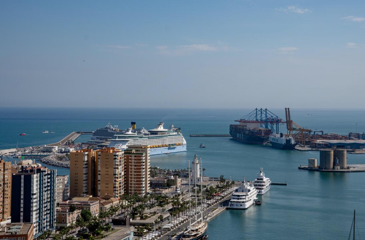 Elevated view of a Mediterranean port with high-rise buildings, a palm-lined promenade, a lighthouse, moored yachts, and a large docked cruise ship.