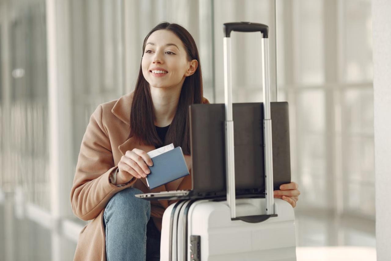 Smiling young woman in a tan coat waiting in an airport terminal, holding a passport with a boarding pass tucked inside and a laptop open on her white suitcase."