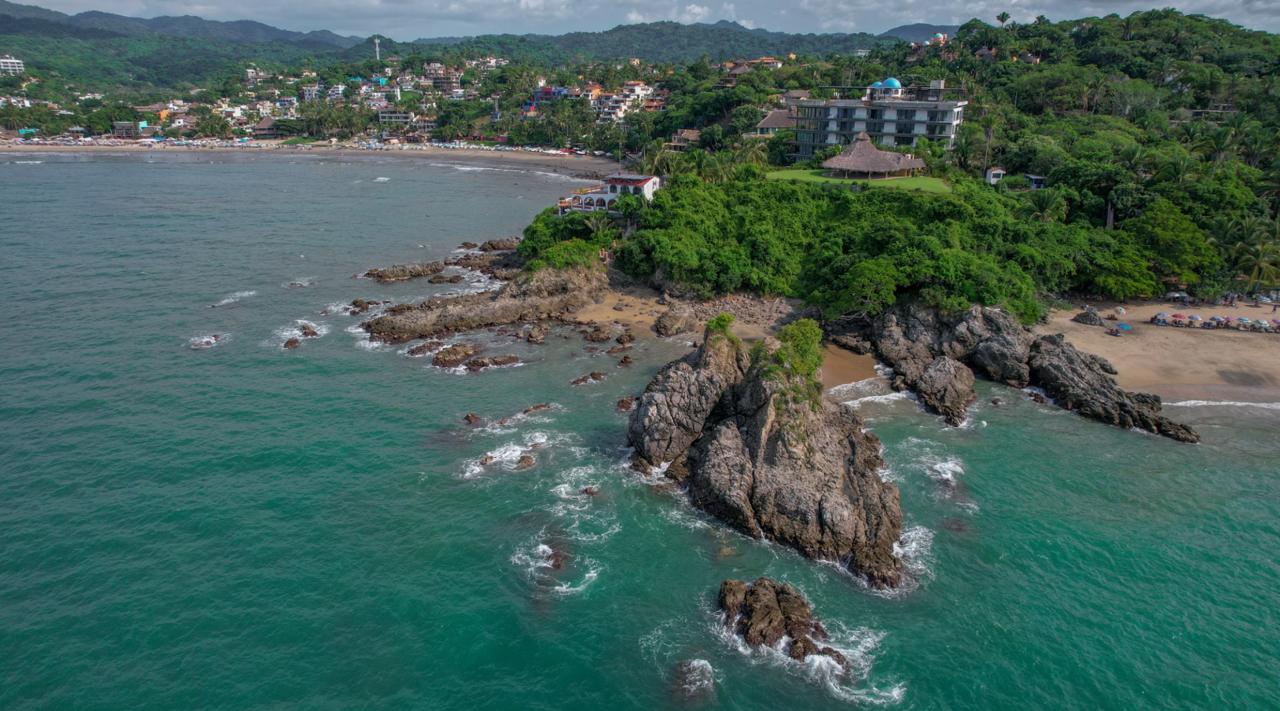 Aerial of the Riviera Nayarit surf town of Sayulita, with a rocky point, an umbrella-dotted beach and forested hills behind.