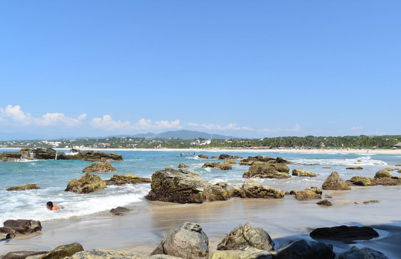 A rocky Pacific shoreline at Puerto Escondido with breaking waves and a swimmer, the town and green hills stretching along the bay behind.