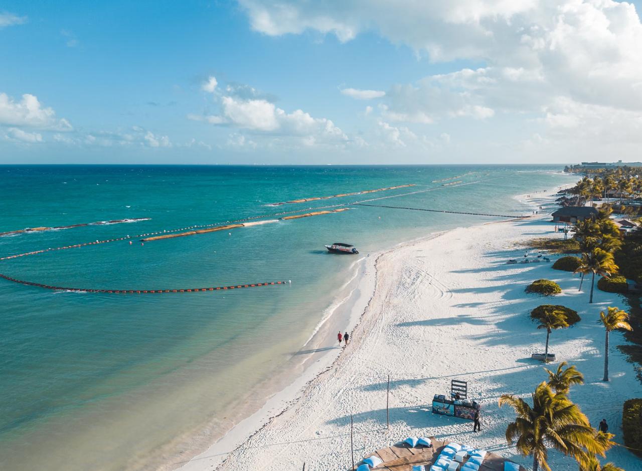 Aerial of a Riviera Maya beach with floating orange sargassum barriers offshore, palm-lined white sand and a resort along the shore.