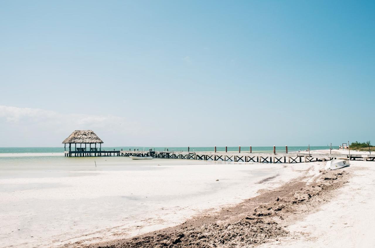 A wooden pier topped with a thatched palapa reaching into the calm shallows off car-free Holbox Island, with white sand and small boats in the foreground.