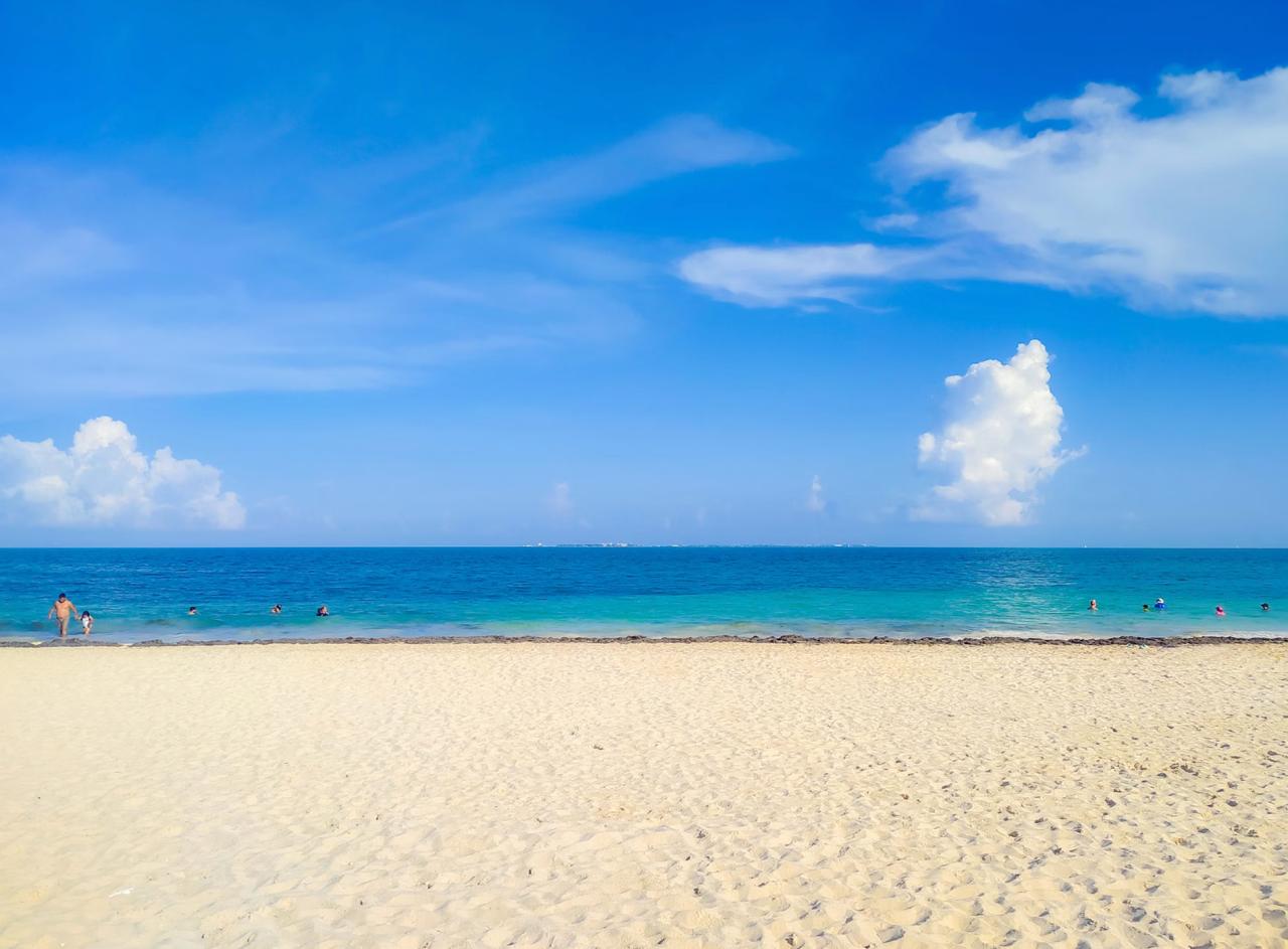 The wide pale-sand public beach at Playa Delfines in Cancún, with swimmers in calm blue Caribbean water under a bright sky.