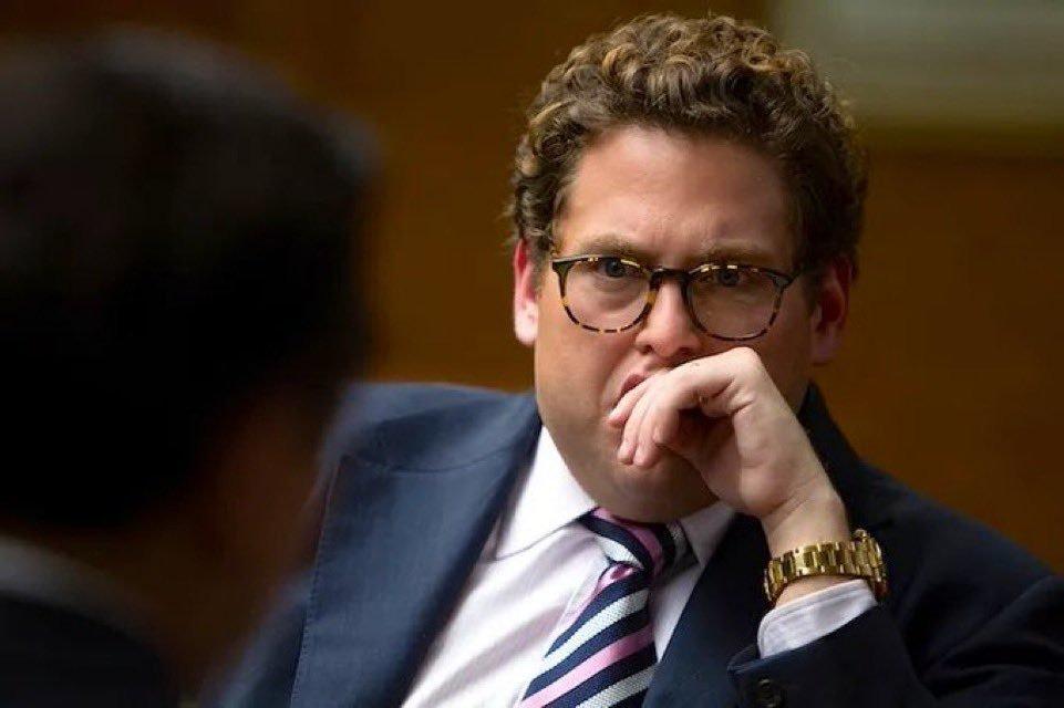 Jonah Hill in glasses resting his chin on his hand looking pensive