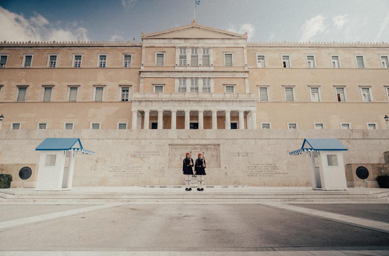 Two Evzones guards in white uniform with pleated kilts marching Parliament ceremony.
