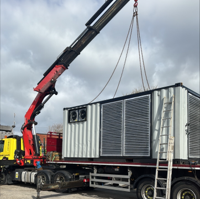 Crane lifting large metal container onto a flatbed truck under a cloudy sky.