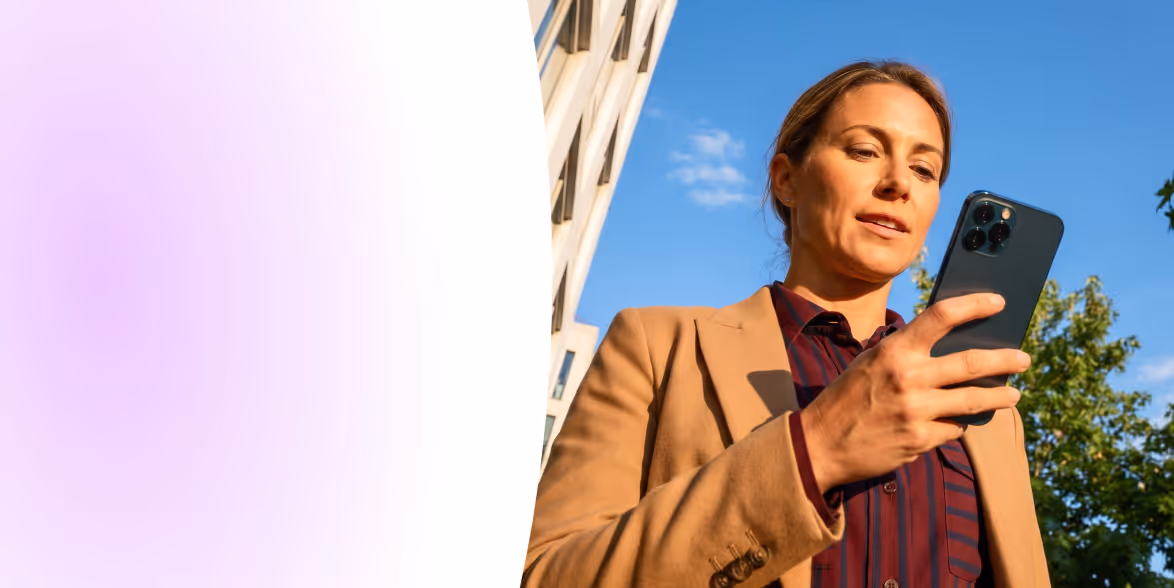 Woman outdoors in blazer looking at her smartphone with a building and trees in the background.